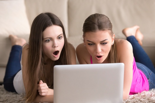 Two cute young women lying on floor in living room, looking with amazement at laptop screen. Shocked teen girlfriends watching movie, surprised by Internet news, impressed by huge online sale discount