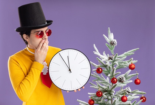 young man in yellow turtleneck and glasses wearing black hat holding wall clock looking at it intrigued standing next to a christmas tree over purple background