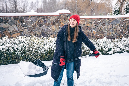 A young woman in a red hat, red mittens and a black jacket cleans snow in the yard in snowy weather.