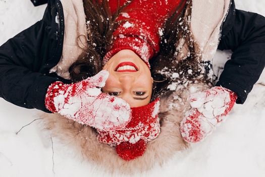 view from above on young pretty candid smiling happy woman in red mittens and knitted hat wearing black coat lying in snow in park, warm clothes, having fun