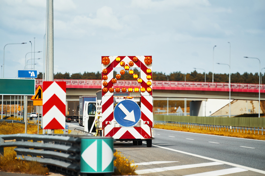 A road works sign with a flashing arrow directs traffic to the different lane on a highway. A service vehicle and a worker are visible behind the sign, working on the road repair. Road works.